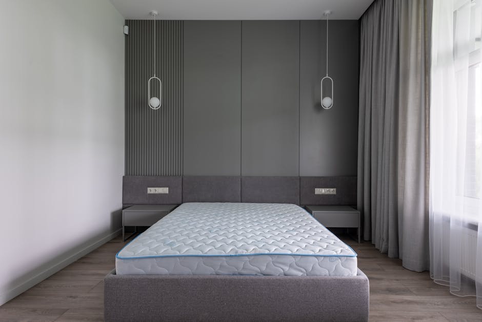 The image displays a modern, minimalistic bedroom with a focus on the bed positioned centrally against a dark grey, textured panelled wall. The bed features a simple, upholstered grey frame and a white mattress with a quilted surface and a subtle wave pattern, resting directly on the frame without bedding or linens. Flanking the bed are two small, matching grey bedside tables with clean, flat surfaces and no visible drawers or handles. Hanging from the ceiling above each side of the bed are contemporary pendant lights with slim, curved white cords and oval-shaped fixtures, contributing to the room's sleek aesthetic. To the right, a large window is covered with sheer white curtains that allow diffuse natural light to fill the space, complemented by a set of heavier grey drapes pulled aside. The flooring consists of light-toned wooden planks, creating a neutral backdrop that emphasizes the room's understated design. The scene suggests a clean, well-maintained interior potentially prepared for temporary or final removal of furnishings, as part of an estate clearance or private disposal, akin to services provided by Waste Removal Merton for rubbish or bulky items like mattresses.