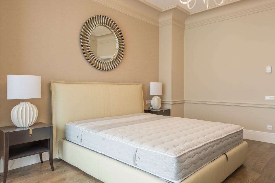 A bedroom interior featuring a large, white, tufted mattress positioned on a cream-colored upholstered bed frame with a padded headboard. The mattress has a textured surface with visible quilting and a slight indentation near the middle. Flanking the bed are two dark wooden bedside tables, each topped with a modern, white ceramic table lamp that has a rounded, ribbed base and a simple cylindrical shade. Above the bed, a round decorative mirror with a thick, textured frame composed of alternating black and silver elements hangs on the beige wall. The room is illuminated softly by natural or artificial light, creating a neutral and calm atmosphere. The walls are decorated with white crown molding and paneling details, contributing to a sophisticated aesthetic. The overall setting suggests an interior space prepared for installation or disposal of household items, in line with private waste handling or rubbish removal services, with the scene representing typical bedroom furnishings within a residential context.
