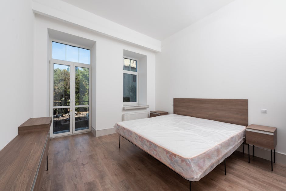 The image displays a modern, minimalistic bedroom with a focus on the bed positioned centrally against a dark grey, textured panelled wall. The bed features a simple, upholstered grey frame and a white mattress with a quilted surface and a subtle wave pattern, resting directly on the frame without bedding or linens. Flanking the bed are two small, matching grey bedside tables with clean, flat surfaces and no visible drawers or handles. Hanging from the ceiling above each side of the bed are contemporary pendant lights with slim, curved white cords and oval-shaped fixtures, contributing to the room's sleek aesthetic. To the right, a large window is covered with sheer white curtains that allow diffuse natural light to fill the space, complemented by a set of heavier grey drapes pulled aside. The flooring consists of light-toned wooden planks, creating a neutral backdrop that emphasizes the room's understated design. The scene suggests a clean, well-maintained interior potentially prepared for temporary or final removal of furnishings, as part of an estate clearance or private disposal, akin to services provided by Waste Removal Merton for rubbish or bulky items like mattresses.