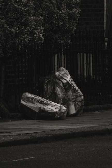 Two large black plastic refuse bags filled with household waste are positioned on the curbside next to a pavement in an outdoor urban setting. The bags appear to be tightly knotted at the top, with some slight creases and folds in the plastic material, which has a slightly glossy finish. They are placed directly on the ground, adjacent to a black metal fence with vertical slats and behind it, visible foliage of dense trees or shrubs with dark leaves, creating a natural background. The scene is illuminated by natural daylight, with minimal shadows indicating overcast or diffuse lighting conditions. The concrete surface of the pavement and the asphalt road are also visible, situating the scene within a typical domestic or semi-private street environment. This type of roadside waste placement might relate to private rubbish collection or on-site clearance services provided by Waste Removal Merton, supporting alternative waste handling methods outside regular council collection schedules, with the focus on efficient rubbish disposal in residential areas.