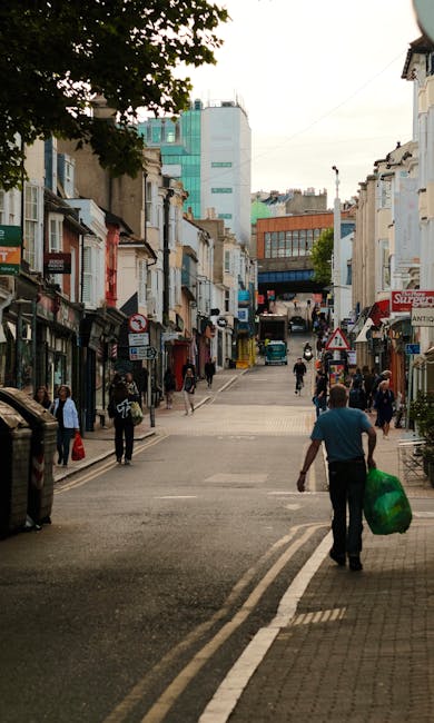 A man walking across a busy urban street with a green plastic bag in his right hand, seen from behind. The street is lined with small shops and commercial buildings on both sides, featuring various signs and awnings. Several pedestrians are visible on the sidewalks, some standing and others walking, creating a lively city atmosphere. In the background, taller modern buildings with glass facades and older architectural structures are visible, suggesting a city centre setting. The street surface is paved, with a designated pedestrian crossing area in the foreground. The scene is well-lit with natural daylight, capturing the everyday activity of city life. This setting connects to the context of alternative waste handling and rubbish removal, illustrating the need for efficient collection services like those provided by Waste Removal Merton for urban environments.