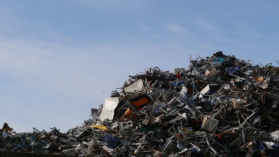 A large mound of assorted scrap metal and discarded household appliances sits outdoors against a clear blue sky. The pile includes broken washing machines, metal frames, twisted wires, and various unidentifiable metal objects, all piled haphazardly with some parts overlapping each other. Visible textures range from smooth, painted surfaces on appliances to jagged, rusted edges of scrap metal. The materials are primarily metallic in colour, with shades of grey, silver, black, and some rust-brown spots. The pile extends across the lower part of the image, occupying the foreground, with no clear boundary, creating a chaotic scene typical of waste management or alternative disposal sites. The bright daylight illuminates the scene evenly, highlighting the metallic sheen and the roughness of the collected debris. This type of setting could relate to the process of off-site rubbish collection or scrap removal, showcasing the kind of waste handled by independent waste removal services such as Waste Removal Merton, which offers rubbish clearance options that differ from standard local authority collections.