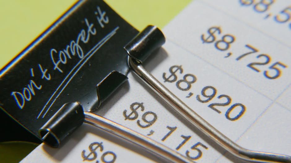 A close-up photograph of a metal binder clip with black plastic handles, clamping onto a printed sheet of paper displaying prices in US dollars with amounts of $59.15, $89.20, and $58.75 visible. The binder clip, positioned in the foreground, has a shiny metallic surface and is partially opened, securing the paper beneath it. The paper appears to be part of a flyer or receipt, with a white background and blue, black, and orange printed text. The scene is well-lit, emphasizing the reflective surface of the metal clip and the clarity of the printed details. The background is slightly blurred, focusing attention on the metal clip and the prices. This image may relate to the concept of organizing or controlling waste management costs, reflecting the importance of managing expenditure in rubbish removal services, as provided by Waste Removal Merton, especially in the context of alternative approaches such as private disposal versus traditional city collection.