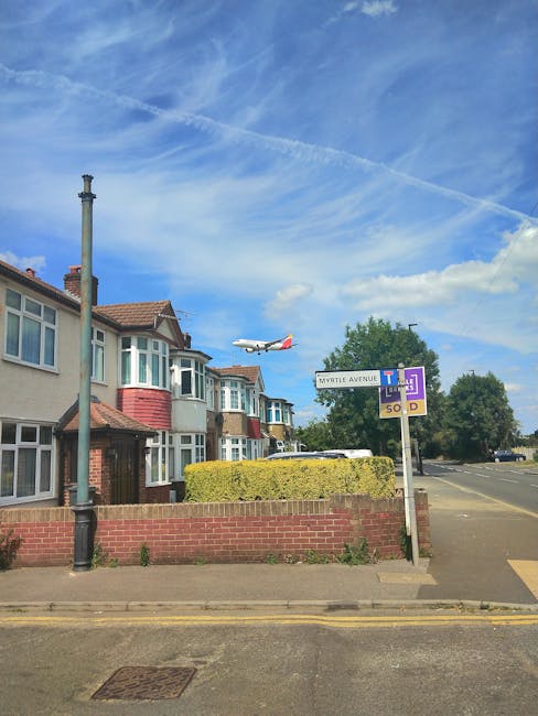 A row of two-story residential houses with bay windows and tiled roofs, situated on a suburban street under a partly cloudy blue sky. The houses are painted in light cream and pastel shades, with some having brickwork details around the entrances. A tall, black lamppost stands on the sidewalk in front of the houses. To the right, there is a street sign indicating the corner of Myrtle Avenue, with a secondary sign beneath it displaying parking restrictions. A neatly trimmed, low hedge runs along the front of the properties, separating the pavement from the private front gardens. An aircraft is visible in the background, flying low across the sky, suggesting proximity to an airport or flight path. The street appears calm, with a few parked cars and an open road extending into the distance. This scene captures a typical residential area where private waste collection or on-site clearance might be relevant, as promoted by waste removal services like Waste Removal Merton, which offers efficient rubbish removal solutions for such neighbourhoods.