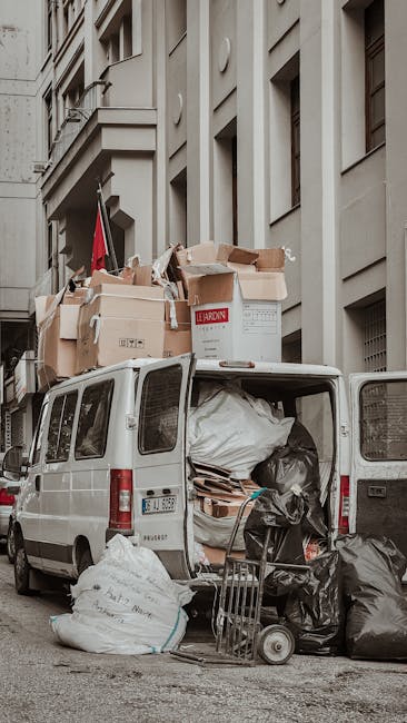 A white panel van parked on a city street with its rear doors open, revealing various types of waste and rubbish inside. The cargo area contains large black and white trash bags, cardboard boxes, and loose items stacked haphazardly. Several cardboard boxes, some with flaps open, are placed on top of the vehicle, including one large box with red text visible on the side. The van is positioned close to a multi-storey building with a beige or light grey facade, featuring architectural details such as columns, niches, and rectangular windows. The scene is outdoors with natural daylight, and the street appears to be in an urban area. Waste removal Merton occasionally handles such on-site clearance and private waste collection, as evidenced by the visible waste accumulation in the vehicle used for rubbish removal, highlighting the type of work involved in managing accumulated household or commercial waste before disposal or recycling processes.