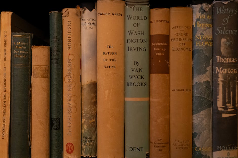 Close-up view of a row of mostly older hardcover books standing upright on a shelf, with visible signs of wear such as torn or faded spines, and some with discolored or peeling covers. The spines display a variety of colors including shades of blue, beige, orange, brown, and black, with titles and authors printed in different fonts and sizes. The textures range from smooth to slightly frayed or damaged, suggesting frequent handling or age. The books are arranged tightly together, with some leaning slightly. The shelf appears to be part of an interior setting, illuminated by soft lighting, highlighting the weathered appearance of the book covers. This image subtly reflects themes related to information, literature, or historical collections that might be managed or cleared during private waste removal or house clearance services such as those offered by Waste Removal Merton while emphasizing the importance of careful handling of valuable or fragile items during such processes.