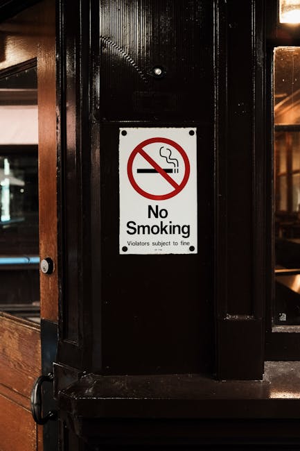 A rectangular white sign with a red prohibition symbol, featuring a lit cigarette emitting smoke, is affixed to a dark, glossy black wooden or metal support beam inside a building. Below the symbol, the sign displays the text 'No Smoking' in bold black letters, with smaller text underneath stating 'Violators subject to fine.' The surrounding environment includes warm-toned wood paneling on the left side and large glass windows or partitions in the background, suggesting an indoor setting such as a restaurant or pub. The lighting is ambient, with a soft glow reflecting off the surfaces, emphasizing the sign's prominence in the scene. This setup relates to waste management by highlighting a non-smoking area, which may be relevant for maintaining hygiene or fire safety standards during rubbish clearance or private disposal activities, often managed by companies like Waste Removal Merton.