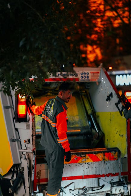 A waste collection worker wearing a high-visibility orange and grey uniform is standing beside a large commercial rubbish truck during dusk or early evening, with the sky showing a fiery orange glow. The worker is positioned to the left of the vehicle, which is painted predominantly white with yellow and red reflective markings, and features various compartments and warning lights. The worker appears to be engaging in a task related to waste collection, possibly adjusting or inspecting the truck, and is holding onto a handle on the side of the vehicle. The scene is set outdoors, possibly on a street or driveway, with a large leafy tree partially obscuring the upper part of the truck. In the background, out of focus, there are additional vehicles or structures, with the overall lighting suggesting the beginning of night, highlighting the necessity of proper waste removal services like those offered by Waste Removal Merton. The detailed description emphasizes the professional setting and the operational aspect of independent rubbish collection or on-site disposal methods.