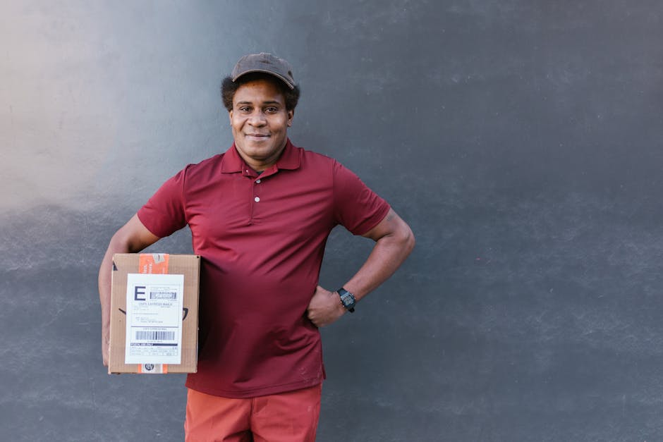 A man of African descent stands outdoors against a plain dark grey wall, holding a small cardboard parcel in his right hand. He is dressed in a maroon collared polo shirt, matching maroon trousers, and a dark-colored cap. His left hand rests on his hip, and he wears a black wristwatch. The parcel has a white shipping label attached, with printed barcodes and text, indicating it is a delivered package. The plain, neutral background emphasizes his neutral expression and casual stance. This scene suggests a typical delivery or courier service, relevant to private waste management practices such as package handling within rubbish removal or collection contexts, indirectly supporting services offered by Waste Removal Merton.