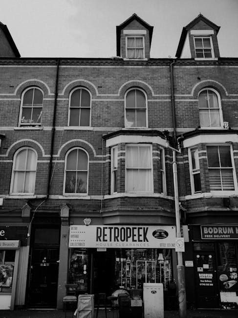 A black and white photograph of a three-story brick building with a retail business named 'Retropeek' on the ground floor, advertising house clearances and collection services. The building features large bay windows on the upper floors, arched window frames on the second floor, and smaller dormer windows in the attic space, with dark roof tiles. In front of the shop, there are two small seats, a bin, and a display stand, set on a pavement area. To the right, part of another storefront with a sign is visible. The overall scene presents an urban street view with the building occupying the central foreground, typical of commercial premises supporting private waste removal services, with the signage suggesting an alternative approach to rubbish collection and disposal. Lighting appears natural with diffuse daylight, and the surroundings are tidy, with no visible rubbish or debris.