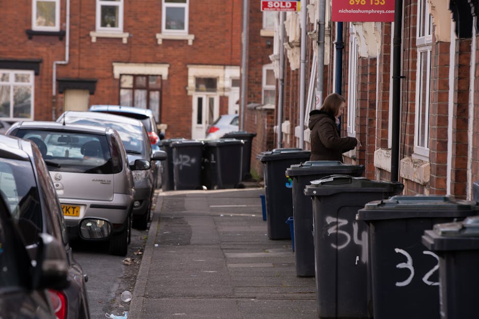 A residential street scene featuring multiple black wheelie bins lined up along the sidewalk adjacent to a row of red brick terraced houses. The bins are made of plastic with hinged lids, some of which are slightly open, revealing black or dark-colored waste inside. A woman wearing a brown coat and hood is seen near the bins, seemingly engaging in private waste disposal or rubbish collection activity. The pavement is concrete with some scattered debris, and the street is narrow with parked cars, including a silver hatchback in the foreground, occupying most of the available space. In the background, additional waste bins are visible, along with a brick building featuring a 'Sold' sign and dotted with white window frames. The overall scene is lit with natural daylight, providing a clear view of waste receptacles ready for collection, aligned with waste removal services focused on independent or private disposal methods.