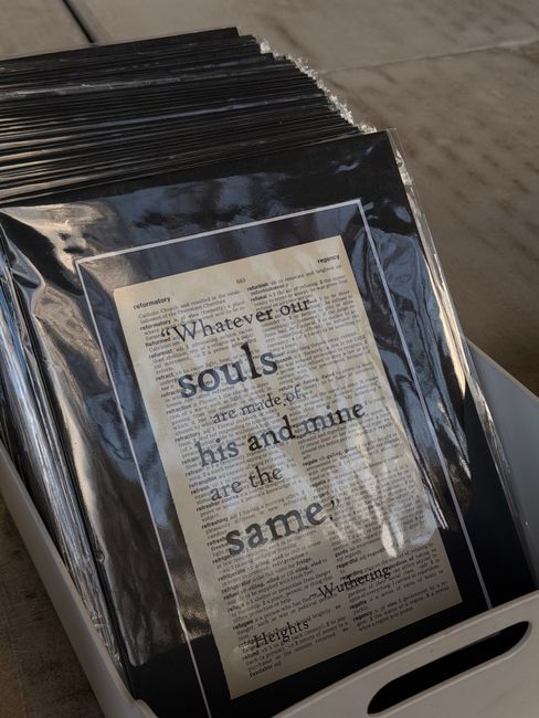 A clear plastic sleeve containing a printed document is positioned on top of a stack of black plastic-covered items. The stack appears to be placed inside a white container with a cut-out handle on the right side. The plastic sleeves have a glossy finish, reflecting light, and show text including the phrase 'Whatever our souls are made of, his and mine are the same,' with additional, smaller text visible beneath. The background includes a concrete or paved surface, suggesting an outdoor or warehouse environment. The scene relates to private or alternative waste handling, consistent with rubbish removal services, and the items in the image are likely retained for disposal or collection by Waste Removal Merton, aligned with their same-day rubbish removal offerings in SW19.