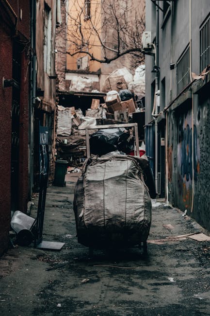 A residential street scene featuring multiple black wheelie bins lined up along the sidewalk adjacent to a row of red brick terraced houses. The bins are made of plastic with hinged lids, some of which are slightly open, revealing black or dark-colored waste inside. A woman wearing a brown coat and hood is seen near the bins, seemingly engaging in private waste disposal or rubbish collection activity. The pavement is concrete with some scattered debris, and the street is narrow with parked cars, including a silver hatchback in the foreground, occupying most of the available space. In the background, additional waste bins are visible, along with a brick building featuring a 'Sold' sign and dotted with white window frames. The overall scene is lit with natural daylight, providing a clear view of waste receptacles ready for collection, aligned with waste removal services focused on independent or private disposal methods.