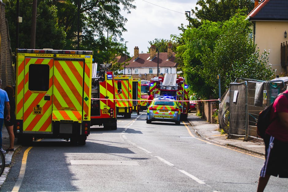A scene on a residential street during an emergency response featuring multiple emergency vehicles including a London Ambulance and police cars with blue lights activated. The vehicles are positioned across the road, partially blocking the street, with the ambulance at the forefront displaying high-visibility yellow and red chevron markings on the rear. Behind the emergency vehicles, several fire trucks are arranged in a line, their bright yellow and red livery standing out against the greenery of nearby trees and bushes. To the right, a makeshift barrier made of fencing material is set up, possibly to control access or secure the area. Pedestrians are visible on either side of the street, some observing the scene while others walk past. The backdrop includes a row of houses with pitched roofs and chimneys, characteristic of a suburban neighbourhood, under partly cloudy skies. This setup, with emergency services present, suggests an incident requiring coordinated response and possible private or non-local-authority involvement in managing the scene, akin to alternative waste handling or on-site clearance operations facilitated by specialised teams or services like Waste Removal Merton.