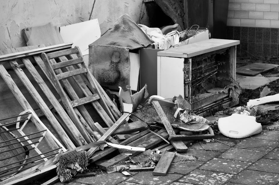 A cluttered outdoor scene showing a mass of discarded wooden furniture including a leaning wooden bed frame and slatted items, alongside a damaged and partially overturned white kitchen cabinet with visible black grime and corrosion on its surface. Surrounding debris includes broken pieces of wood, plastic waste such as a white toilet seat, and miscellaneous objects scattered over a paved surface. The background features a brick wall and a wooden fence, suggesting the area is part of a backyard or garden space, now used for dumping unwanted rubbish. The overall atmosphere indicates an area awaiting collection or clearance, typical of private waste disposal services like those offered by Waste Removal Merton, with visible signs of neglect and accumulated refuse for removal in a professional rubbish removal context.