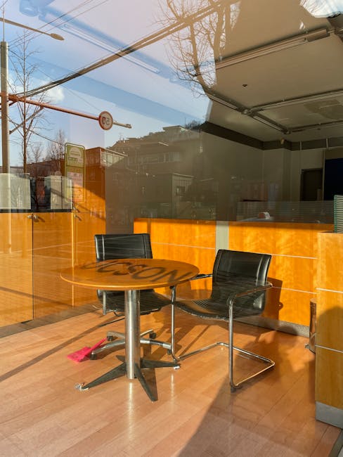 Inside a modern office space with large window panels reflecting the street outside, including a bus stop and street signage. The room features a round wooden table with a polished surface and a sturdy metal pedestal base, accompanied by two black plastic chairs with metal frames, positioned on a light wood laminate floor. Behind the table, an orange reception counter with a smooth finish extends along the wall, partially visible through the glass. The ceiling has built-in lighting fixtures, and the overall environment appears clean and organized, typical of a commercial or client-facing area. The window allows natural daylight to illuminate the interior, creating a bright and open atmosphere suitable for meetings or casual discussions. This setting aligns with a professional business environment that might utilize external waste or rubbish removal services, facilitated by companies like Waste Removal Merton, to maintain cleanliness and operational standards.
