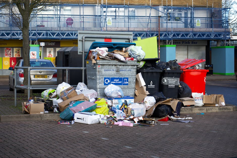 A collection of overflowing rubbish bins and numerous scattered waste items on a paved parking area in front of a commercial building. The waste includes cardboard boxes, plastic bags, paper, and packaging materials, some spilling onto the ground. The central grey bin is filled with assorted papers, cardboard, and plastic, with its lid open and additional rubbish leaning against it. To the right, there are black and red rubbish bins, also overflowing, with black plastic trash bags and loose waste around them. In the background, a parked grey car is visible on the left, and part of a building with blue scaffolding and a shop frontage with signage can be seen behind the rubbish, indicating a busy urban setting. The scene suggests a situation where waste has not been collected recently, highlighting the need for proper rubbish removal services, such as those offered by Waste Removal Merton, to manage bulky waste and debris effectively, especially in private or alternative disposal contexts.