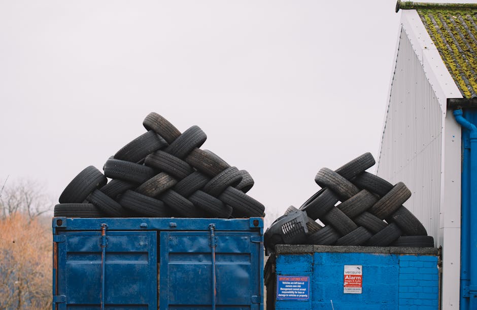 The image shows two blue skip containers filled with used black rubber tires, stacked in an outdoor setting next to a corrugated metal building with a moss-covered roof. The larger container on the left is filled with tires piled high, some leaning randomly while others are stacked more neatly, with visible details such as tread patterns and weathered surfaces. The smaller container on the right also contains a number of tires arranged in a similar fashion, with a few tires protruding over the edge. The background features an overcast sky, slightly blurred trees, and a portion of the building's white wall with a blue drainage pipe. The scene appears to be part of a waste disposal or tyre recycling operation, and the containers are used for bulk storage of discarded tires. This setup may relate to private waste management or independent collection services, exemplifying the typical environment for tyre and rubber waste storage prior to removal by waste management professionals like Waste Removal Merton.