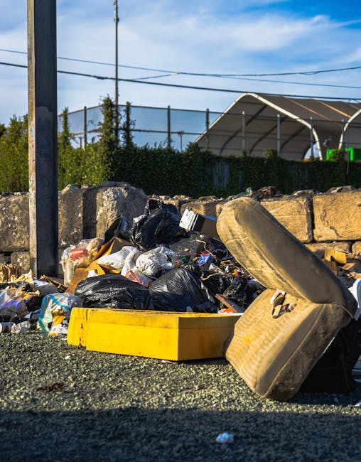 A collection of overflowing rubbish bins and numerous scattered waste items on a paved parking area in front of a commercial building. The waste includes cardboard boxes, plastic bags, paper, and packaging materials, some spilling onto the ground. The central grey bin is filled with assorted papers, cardboard, and plastic, with its lid open and additional rubbish leaning against it. To the right, there are black and red rubbish bins, also overflowing, with black plastic trash bags and loose waste around them. In the background, a parked grey car is visible on the left, and part of a building with blue scaffolding and a shop frontage with signage can be seen behind the rubbish, indicating a busy urban setting. The scene suggests a situation where waste has not been collected recently, highlighting the need for proper rubbish removal services, such as those offered by Waste Removal Merton, to manage bulky waste and debris effectively, especially in private or alternative disposal contexts.